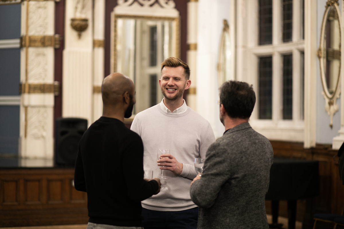 Tall white male in a pale jumper and shirt smiling with a glass of wine whilst talking to two people with their heads to the back of the camera. In a large historial room.