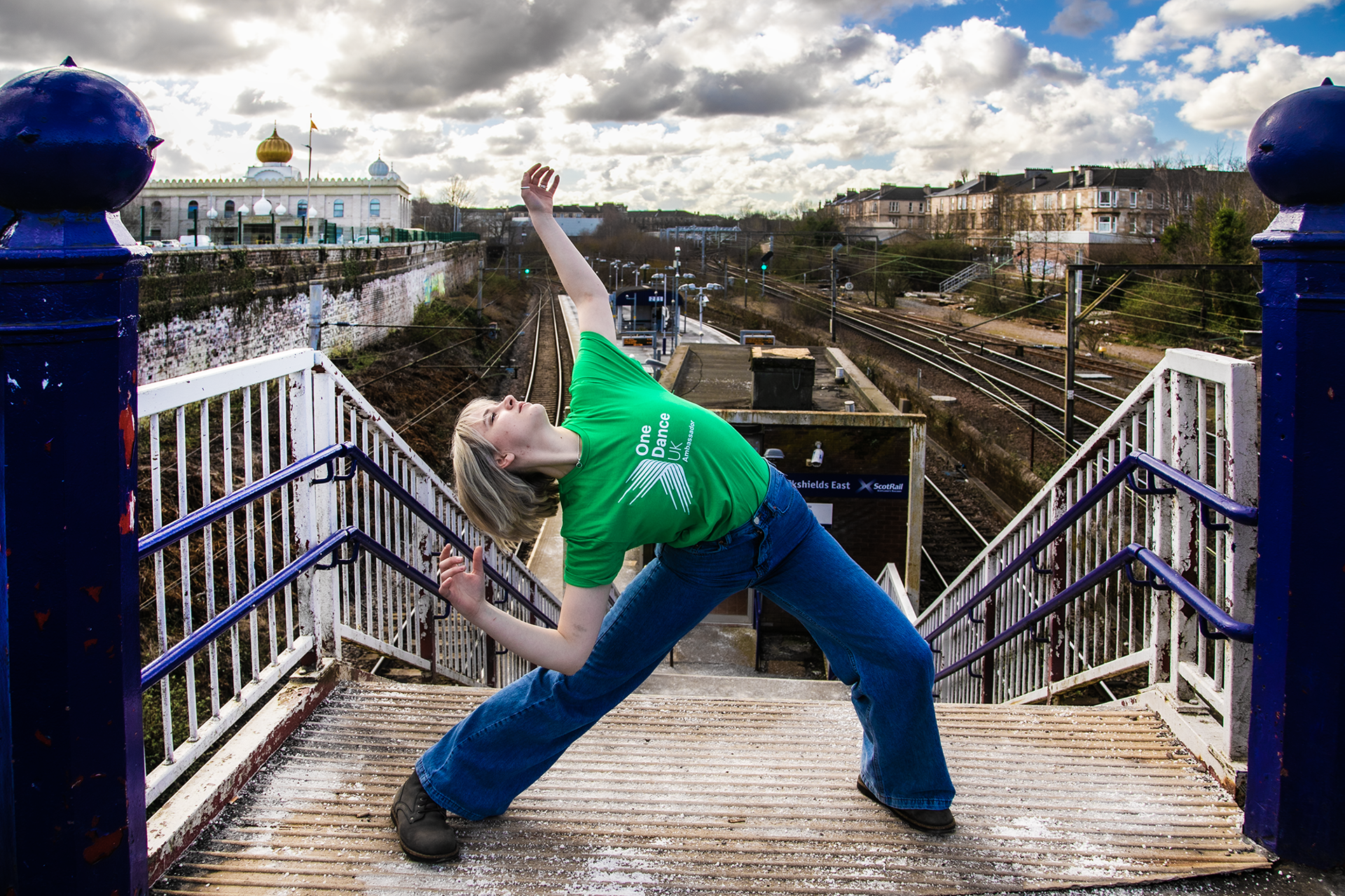 white blonde female lunging with arms high and arched back face pointed to the sky in front of train station platform. Wearing green dance ambassador tshirt and blue jeans white blonde female lunging with arms high and arched back face pointed to the sky in front of train station platform. Wearing green dance ambassador tshirt and blue jeans