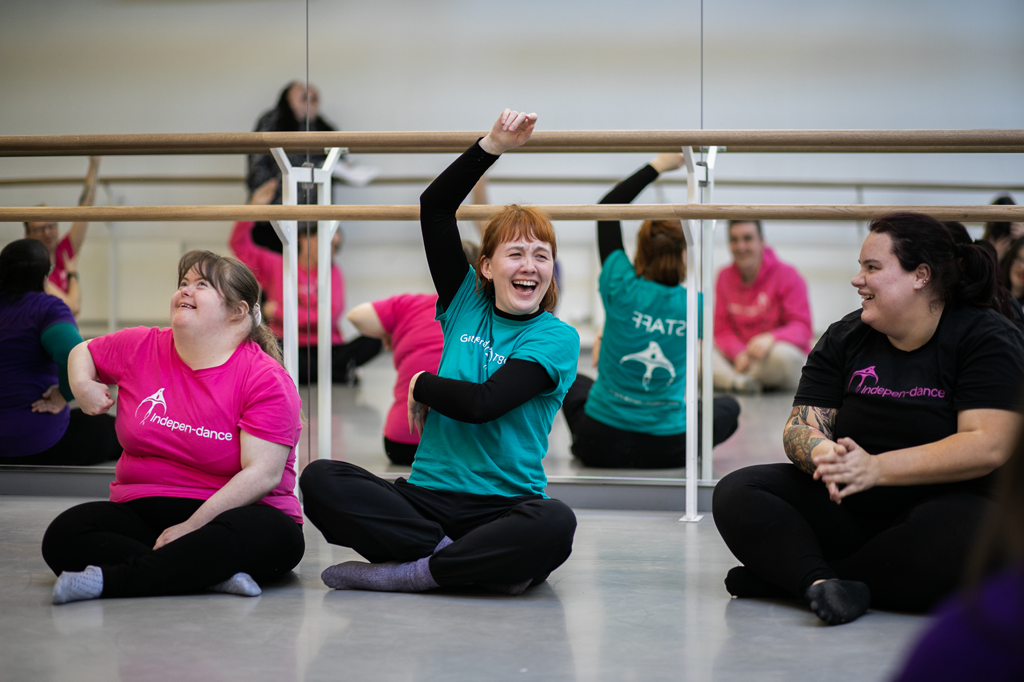 Two white females smiling cross legged, on the left, a disabled dancer in a pink t-shirt, on the right, the group leader raising her arm wearing a teal t-shirt