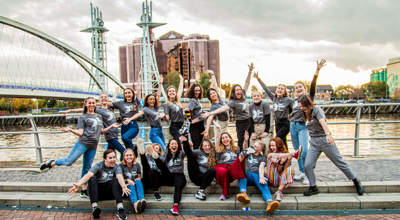 20 male and female dance ambassadors posing in front of Manchester bridge and skyline in the background. Wearing grey Dance ambassador tshirts.