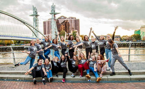 20 male and female dance ambassadors posing in front of Manchester bridge and skyline in the background. Wearing grey Dance ambassador tshirts.