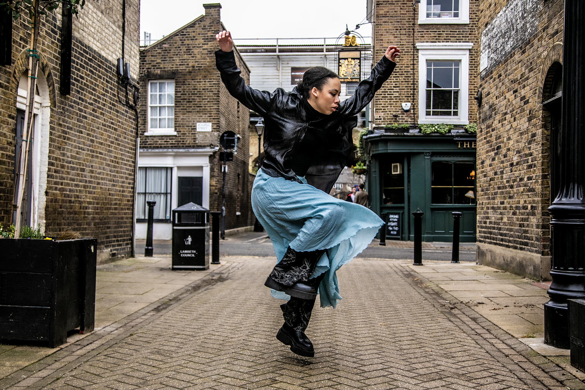 Global majority female dancer skipping down a london street side on with arms in the air . Wearing black leather jacket and blue skirt