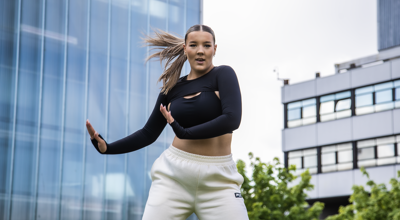 young female dancer with arms to the right side of her infront of Newcastle college building. Wearing black top, white joggers and ponytail swinging to the left