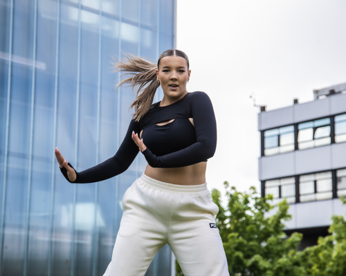 young female dancer with arms to the right side of her infront of Newcastle college building. Wearing black top, white joggers and ponytail swinging to the left