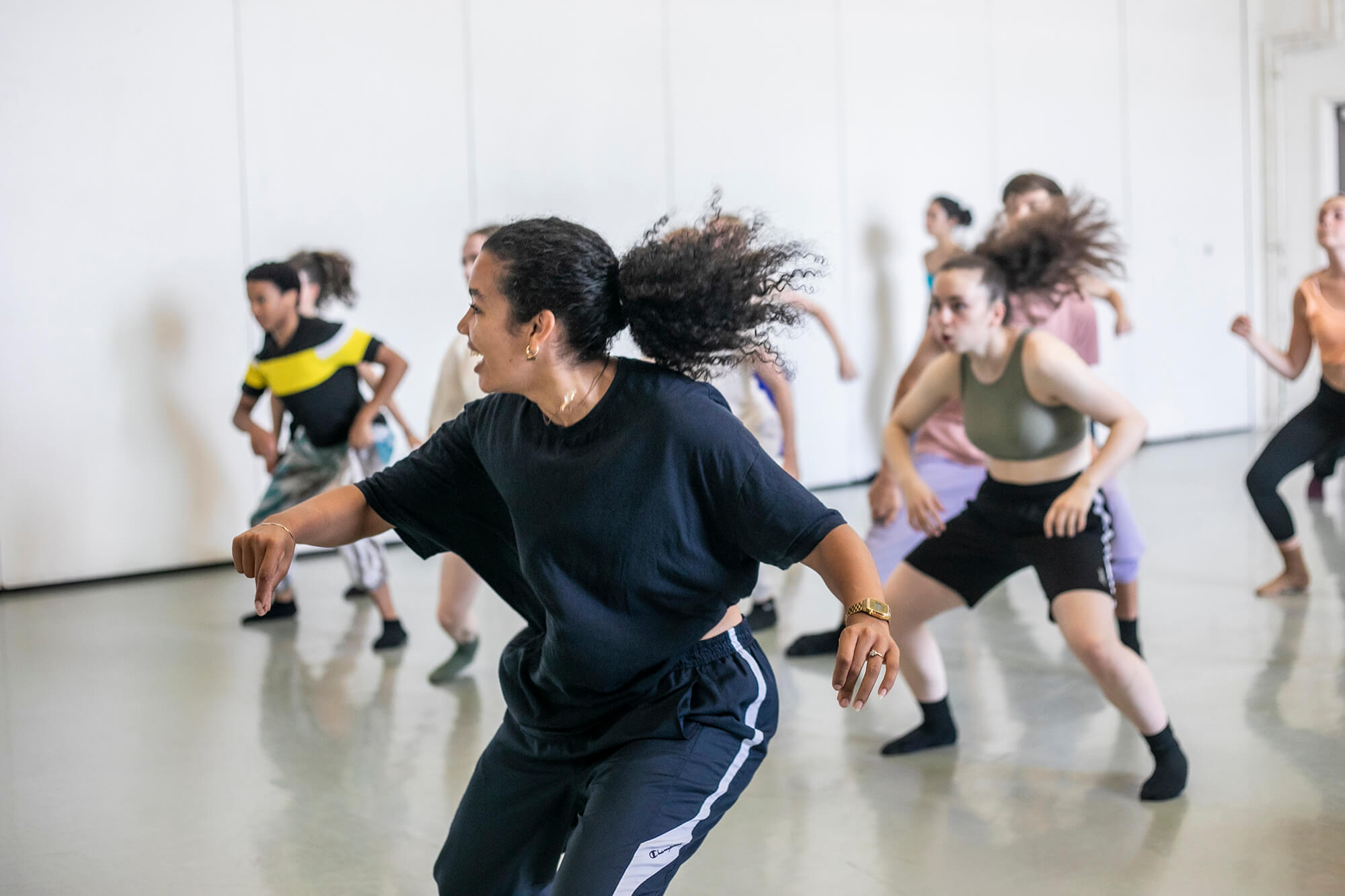 Female global majority dancer with curly hair flicked in a pony tail teaching a hip hop class. students copying the movement behind all wearing bright colours in dance studio