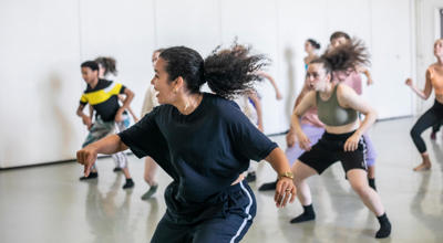 Female global majority dancer with curly hair flicked in a pony tail teaching a hip hop class. students copying the movement behind all wearing bright colours in dance studio