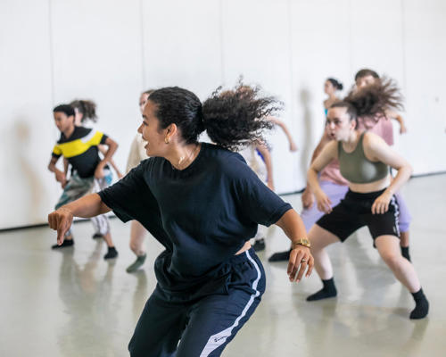 Female global majority dancer with curly hair flicked in a pony tail teaching a hip hop class. students copying the movement behind all wearing bright colours in dance studio