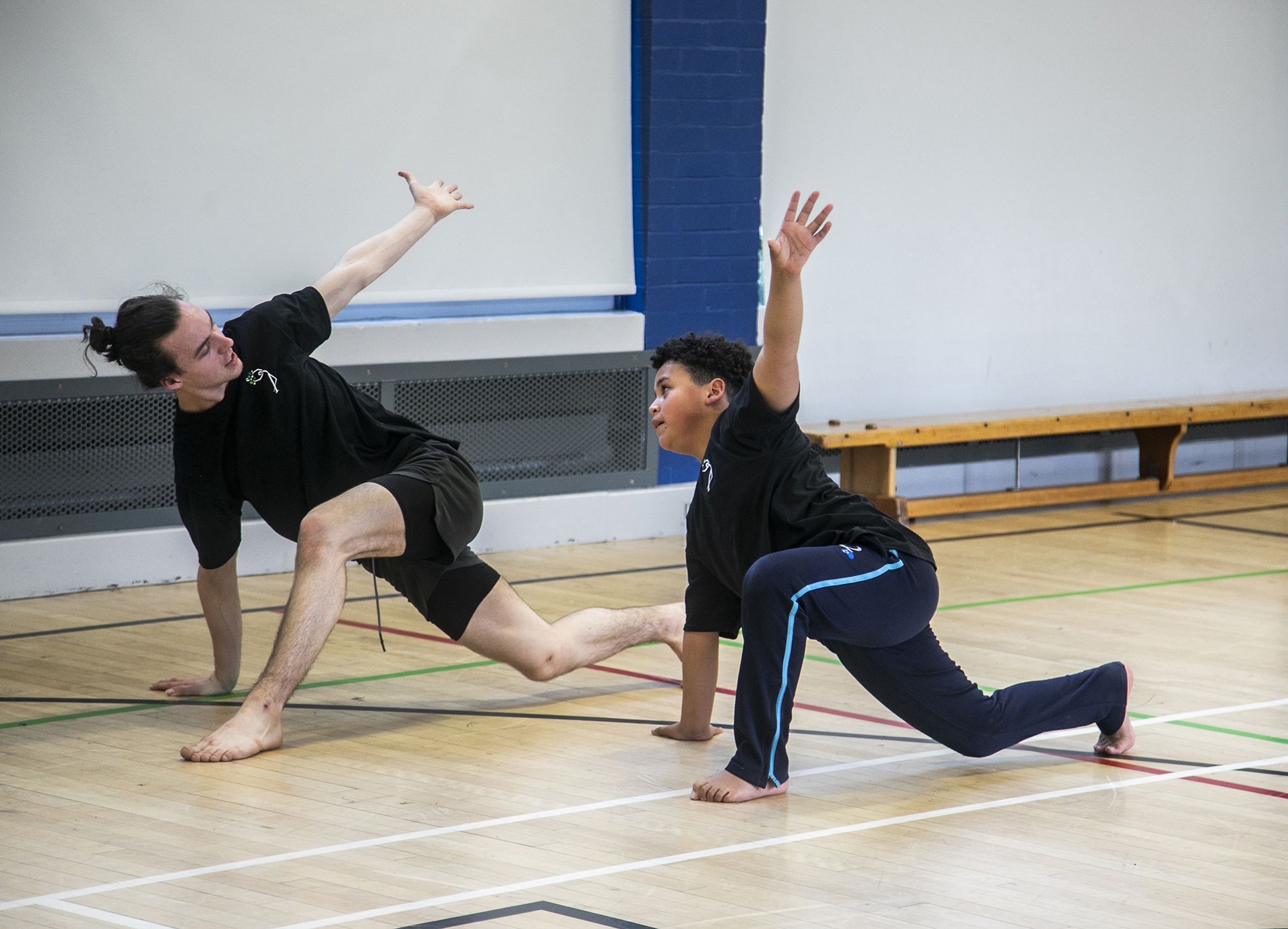 teenage male dance teacher lunging with one arm in the air teaching young boy dancer lunging with the mirrored arm in the air. In sports hall both wearing black PE kit teenage male dance teacher lunging with one arm in the air teaching young boy dancer lunging with the mirrored arm in the air. In sports hall both wearing black PE kit