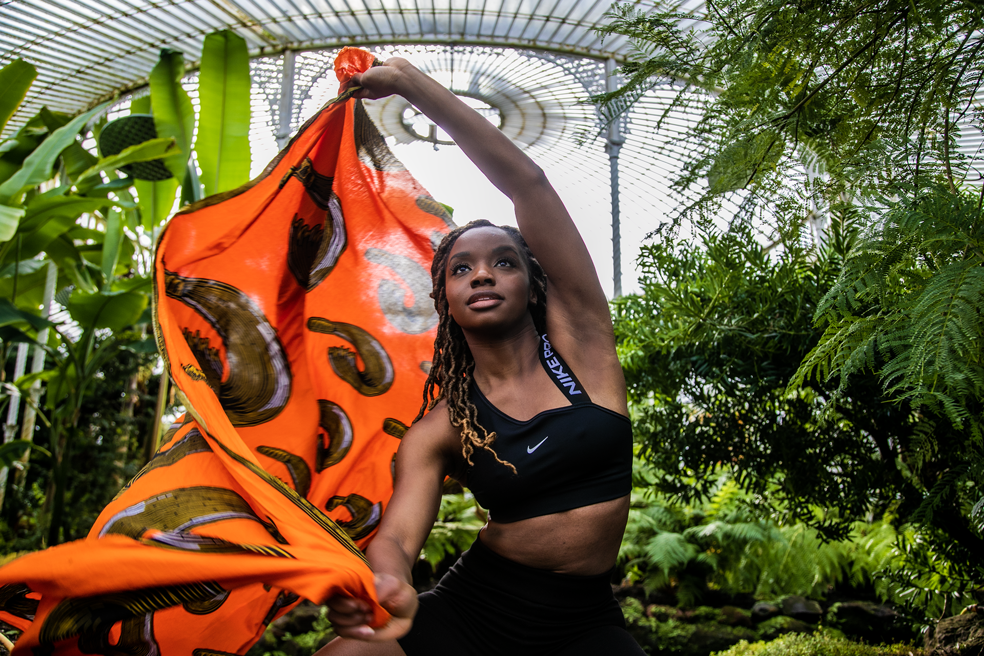 Black female dancer with long brown braids moving a bright orange scarf in the air, looking above the camera, in greenhouse surrounded by plants. Wearing black crop top and black cycling shorts.  