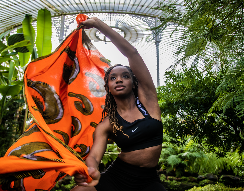 Black female dancer with long brown braids moving a bright orange scarf in the air, looking above the camera, in greenhouse surrounded by plants. Wearing black crop top and black cycling shorts.  