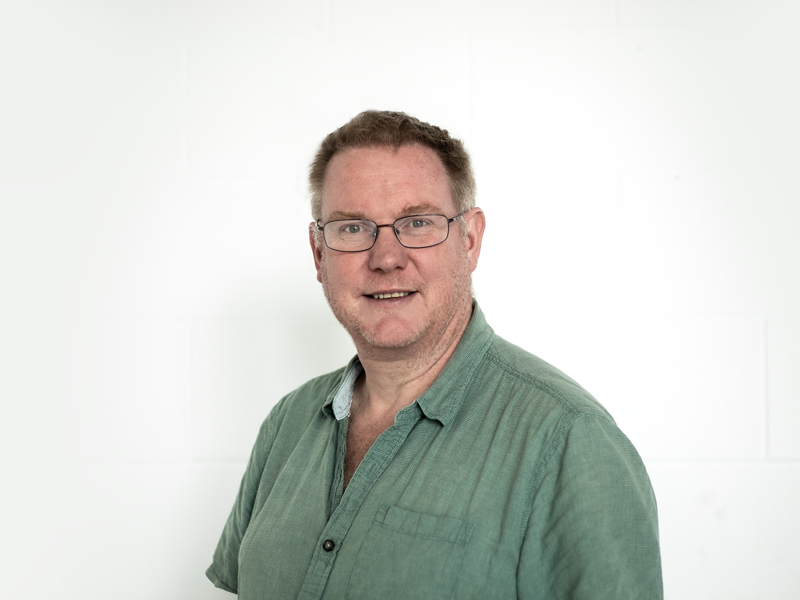 Headshot of Paul Hibbert. White light haired  male smiling at the camera wearing glasses and a green shirt