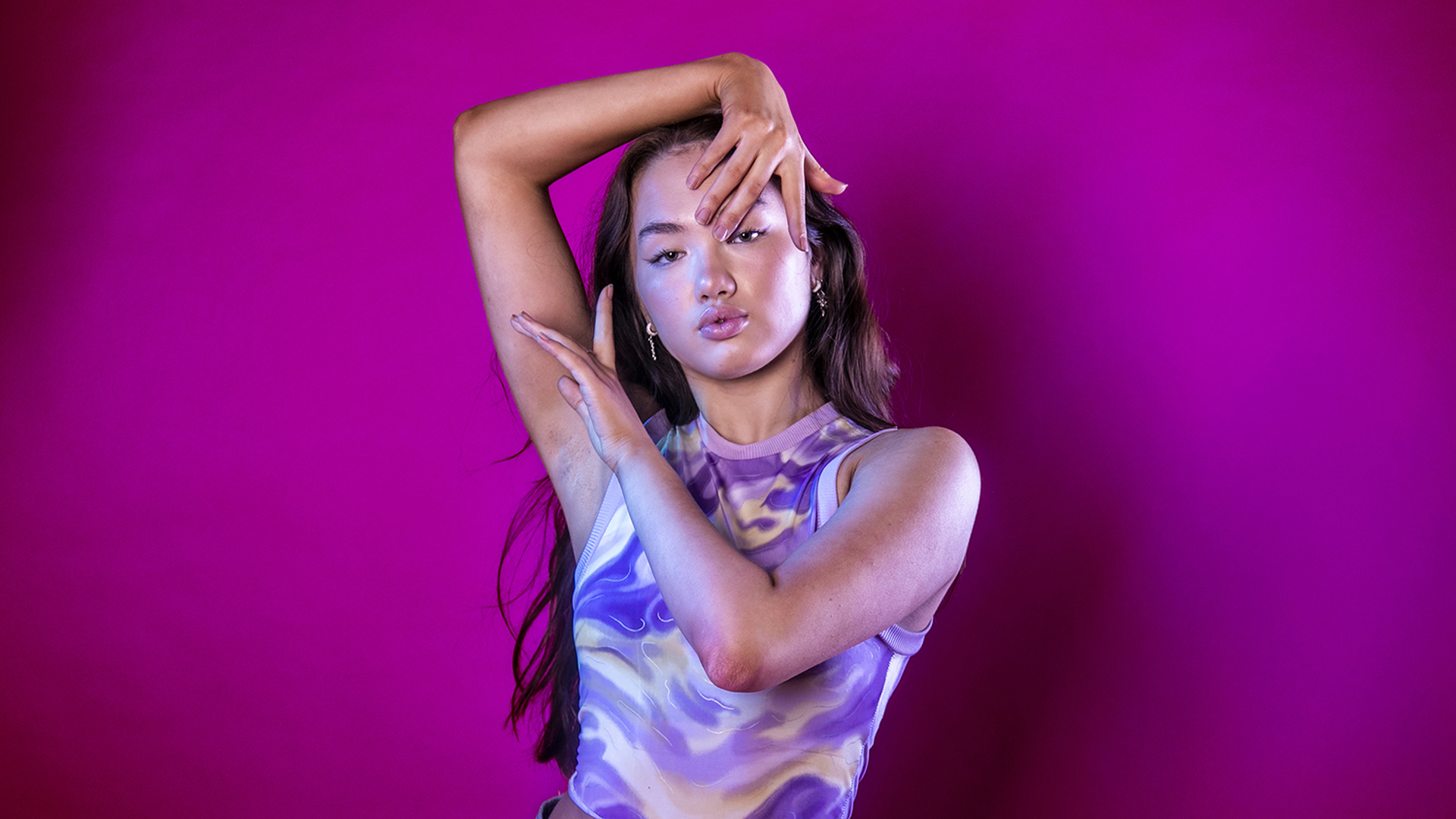 global majority dancer with hands infront and around face on hot pink studio background. Wearing marbled purple crop top.