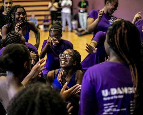 group of young black female dancers hundled together clapping dancing and cheering wearing purple tshirts in a dance studio 