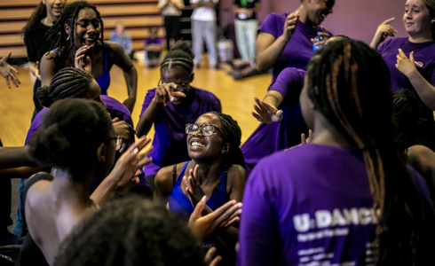 group of young black female dancers hundled together clapping dancing and cheering wearing purple tshirts in a dance studio 