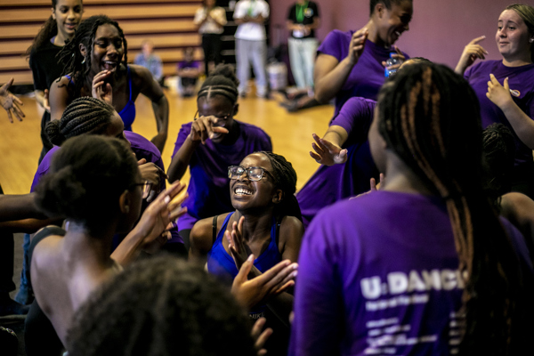 group of young black female dancers hundled together clapping dancing and cheering wearing purple tshirts in a dance studio 