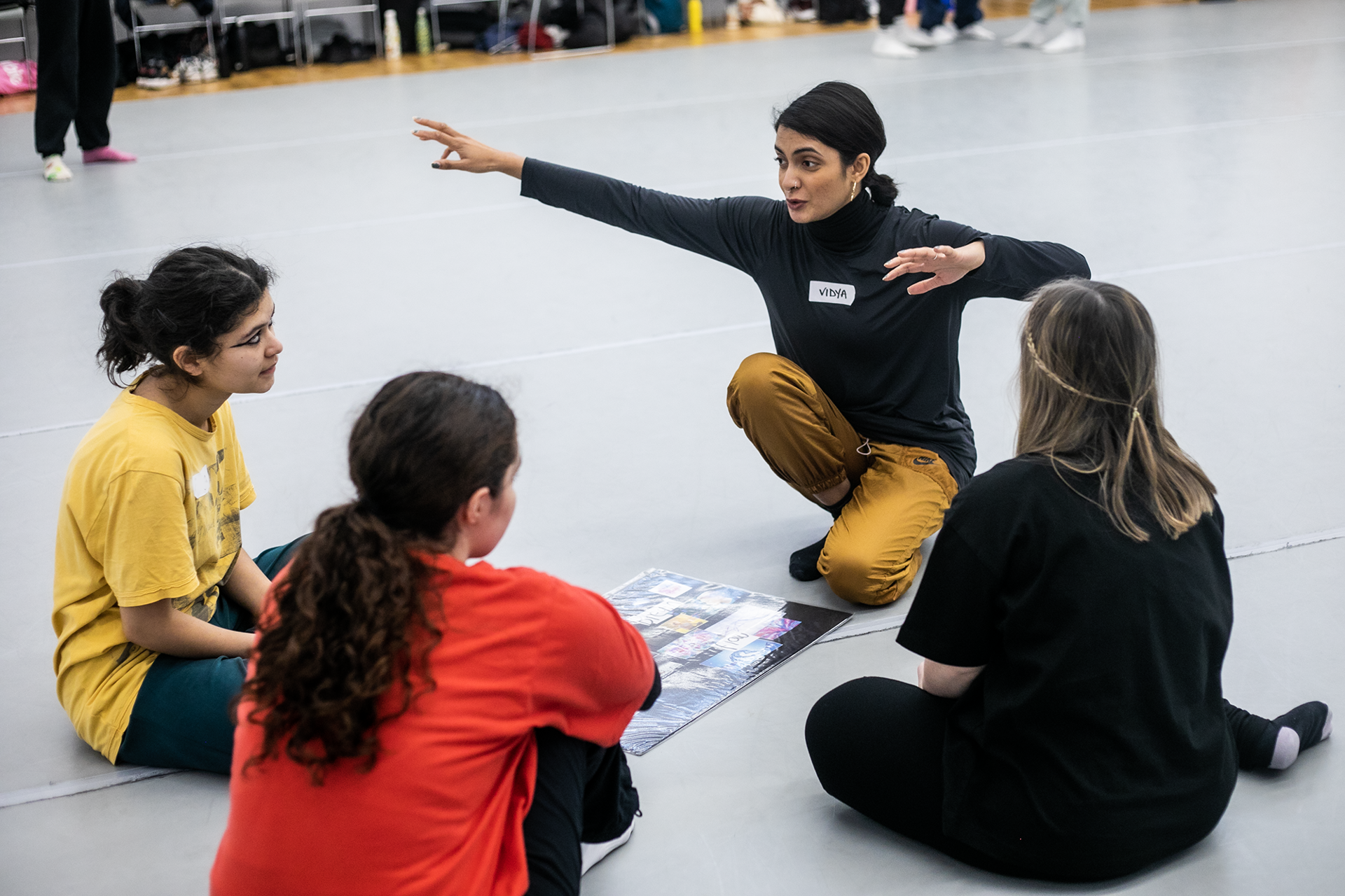 Global majority dance teaching kneeling down with 4 young female dance students gesturing with arms out wide with a mood board in front of them in a dance studio. Global majority dance teaching kneeling down with 4 young female dance students gesturing with arms out wide with a mood board in front of them in a dance studio.