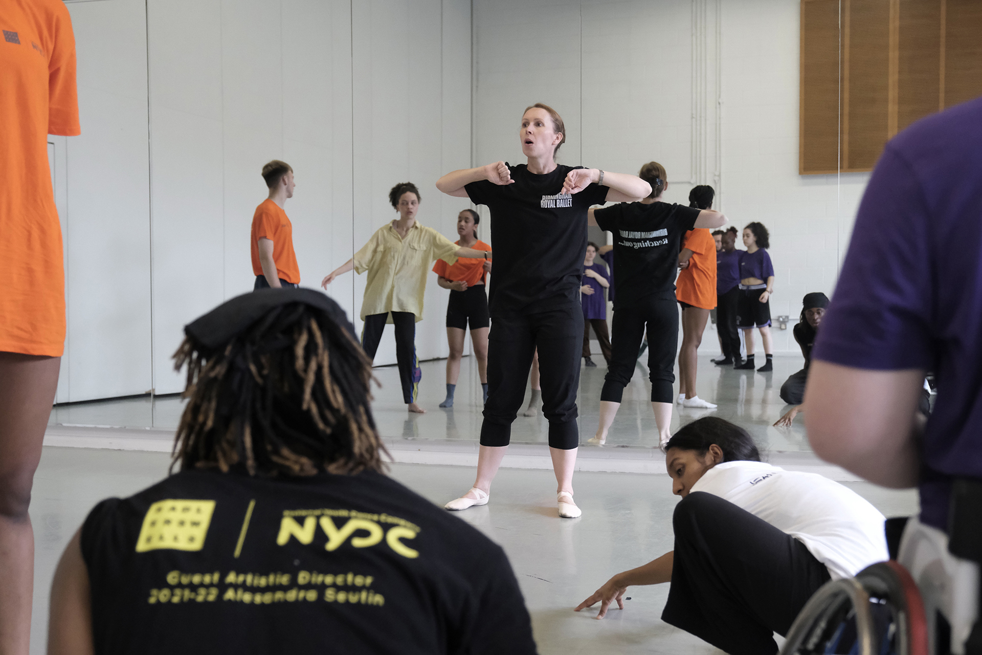 white female dance teacher guestering to a room full of students who are wearing brightly coloured clothes with a mirrored background. In Dance studio. 
