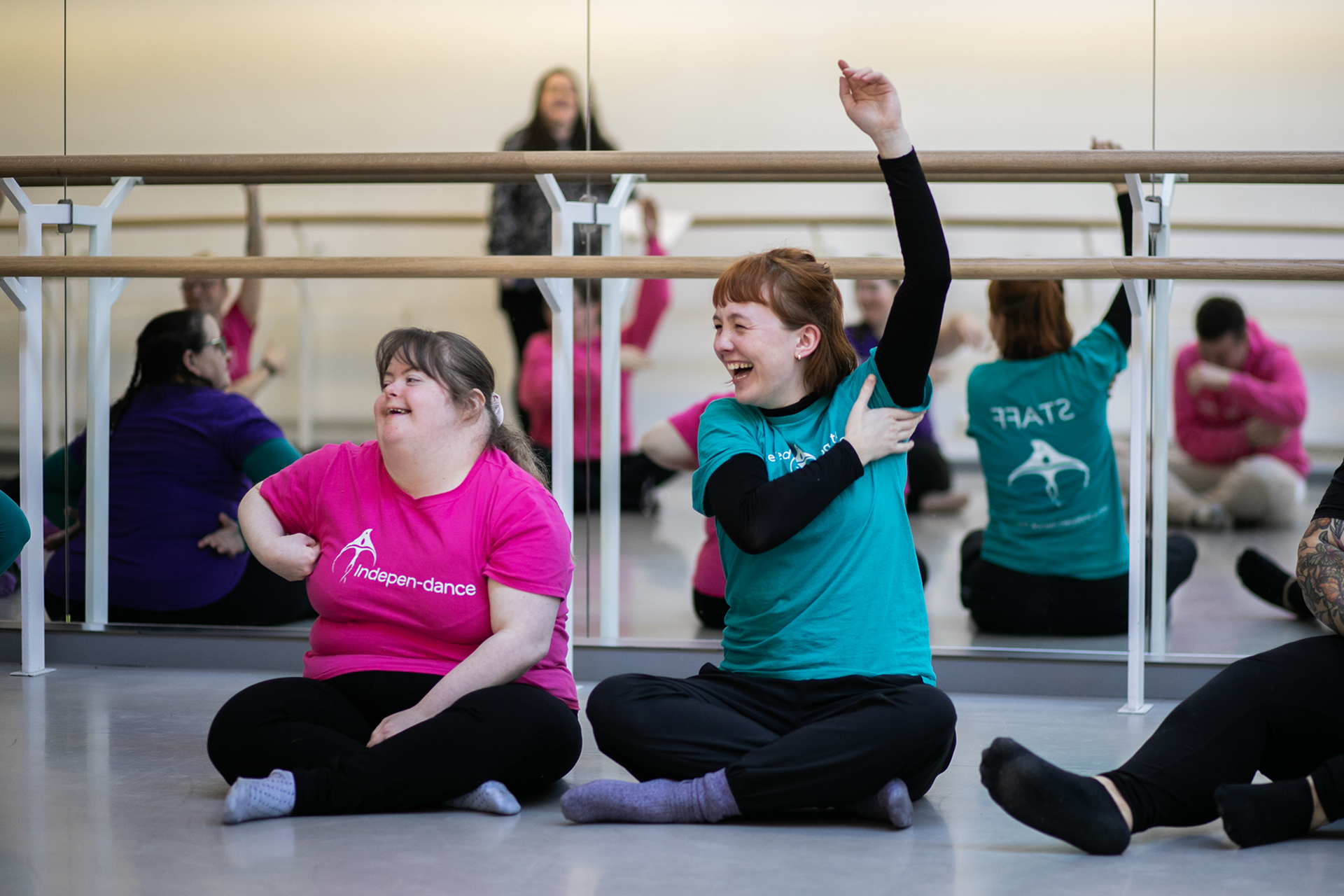 Two white females smiling cross legged, on the left, a disabled dancer in a pink t-shirt, on the right, the group leader raising her arm wearing a teal t-shirt