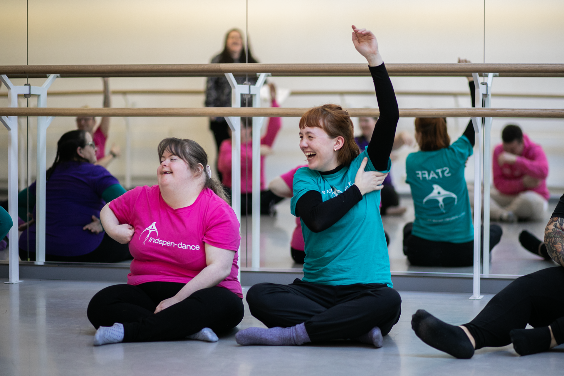 Two white females smiling cross legged, on the left, a disabled dancer in a pink t-shirt, on the right, the group leader raising her arm wearing a teal t-shirt Two white females smiling cross legged, on the left, a disabled dancer in a pink t-shirt, on the right, the group leader raising her arm wearing a teal t-shirt