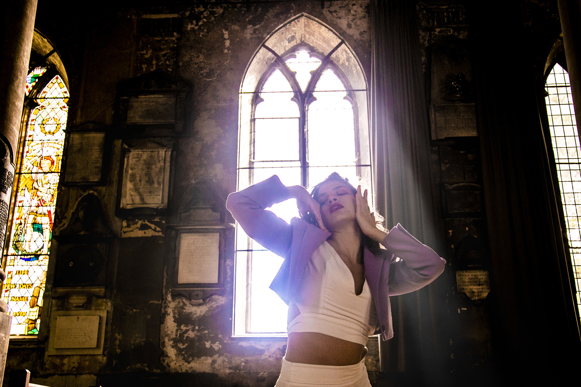 female dancer with hands around the face backlit by church window. Wearing white jumpsuit and purple jacket