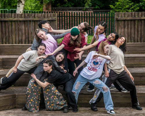 Eleven young male and female dancers huddled in a group on wooden steps moving around eachother with stretched arms. Causually dressed. 