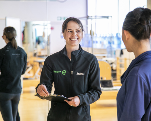 White female with brown tied back hair wearing black zip up jumper holding a clipboard and smiling whilst looking at a white female with tied up black hair wearing purple jumper whos face is turned away from the camera. With mirrored background in a fitness setting.