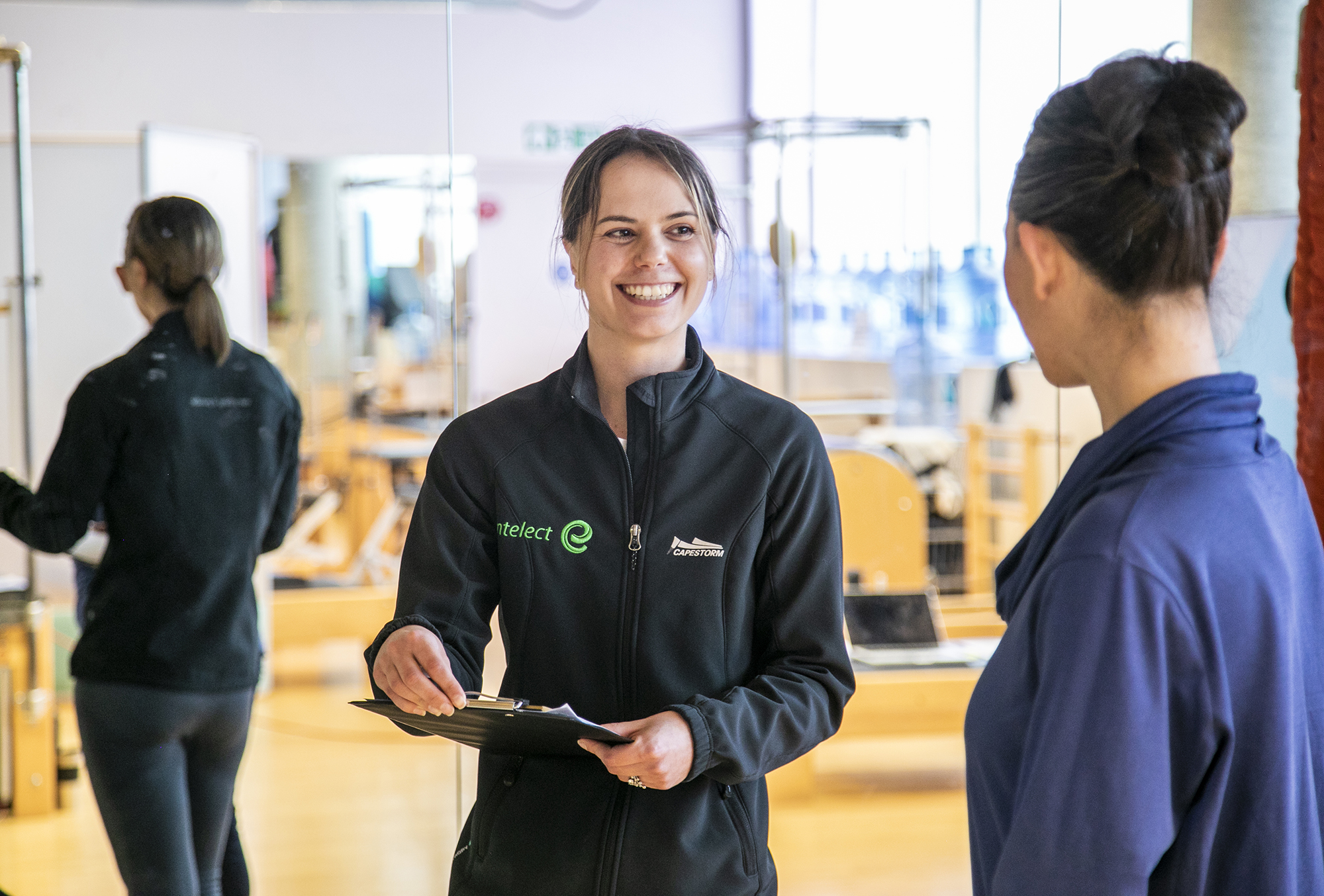 White female with brown tied back hair wearing black zip up jumper holding a clipboard and smiling whilst looking at a white female with tied up black hair wearing purple jumper whos face is turned away from the camera. With mirrored background in a fitness setting.