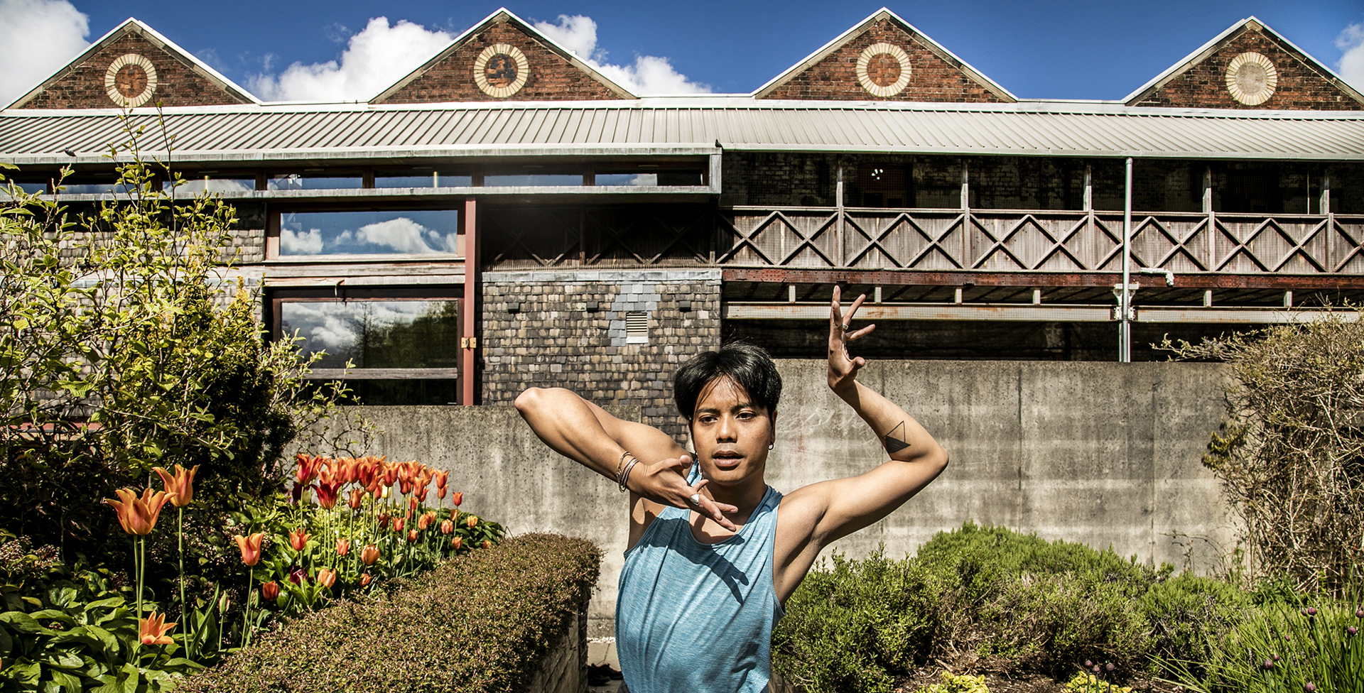Global majority male dancer with short black hair, centre of the image with hand framing his face. Wearing blue tank top in front of tulips and triangular shaped building 