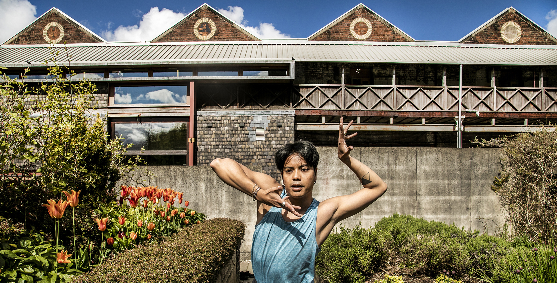 Global majority male dancer with short black hair, centre of the image with hand framing his face. Wearing blue tank top in front of tulips and triangular shaped building Global majority male dancer with short black hair, centre of the image with hand framing his face. Wearing blue tank top in front of tulips and triangular shaped building