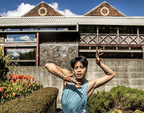 Global majority male dancer with short black hair, centre of the image with hand framing his face. Wearing blue tank top in front of tulips and triangular shaped building 