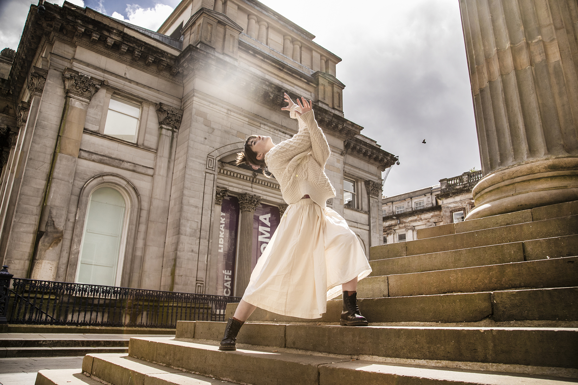 dancer lunging on steps of Glasgow council building with large pillars. White female dancer with brown hair arms interlinked towards the sky. White cable knit jumper and white midi skirt. 