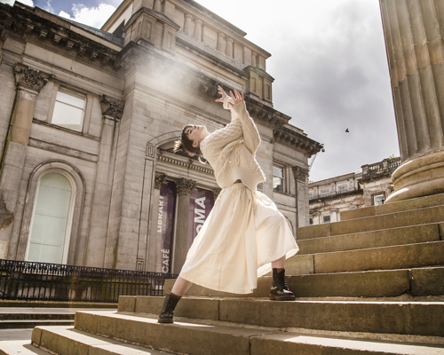 dancer lunging on steps of Glasgow council building with large pillars. White female dancer with brown hair arms interlinked towards the sky. White cable knit jumper and white midi skirt. 