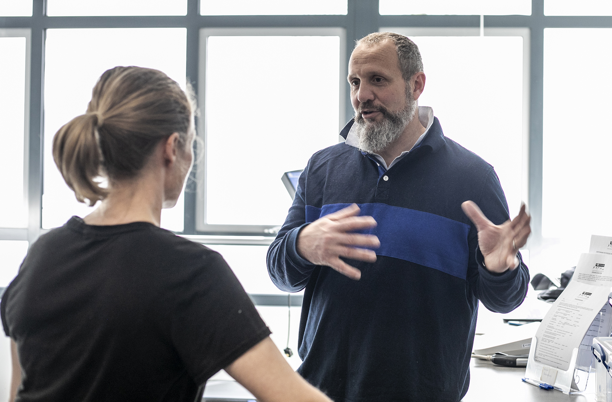 White male physiologist explaining something to white female dancer. In a white laboratory setting with a window in the background