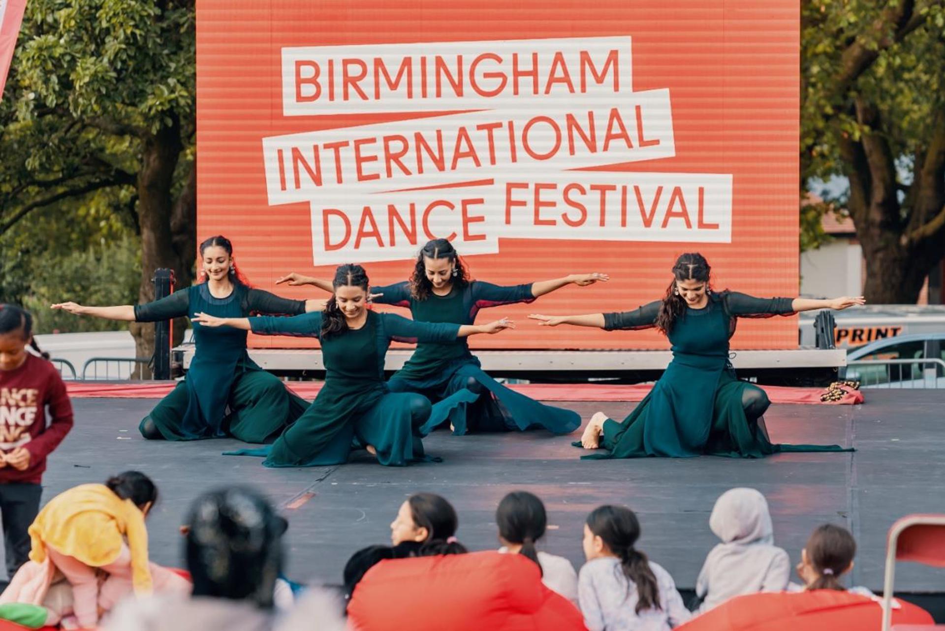 four South Asian dancers all female on one knee with arms out wide in front of large red sign saying Birmingham international dance festival. In front of an outdoor audience.