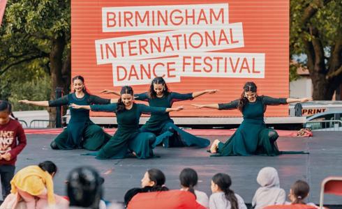 four South Asian dancers all female on one knee with arms out wide in front of large red sign saying Birmingham international dance festival. In front of an outdoor audience.