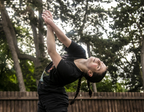 White female dancer with limb difference arms pointing to the top left hand side of the image with arched back. Wearing black t-shirt and two long plaits. In front of soft focus trees White female dancer with limb difference arms pointing to the top left hand side of the image with arched back. Wearing black t-shirt and two long plaits. In front of soft focus trees