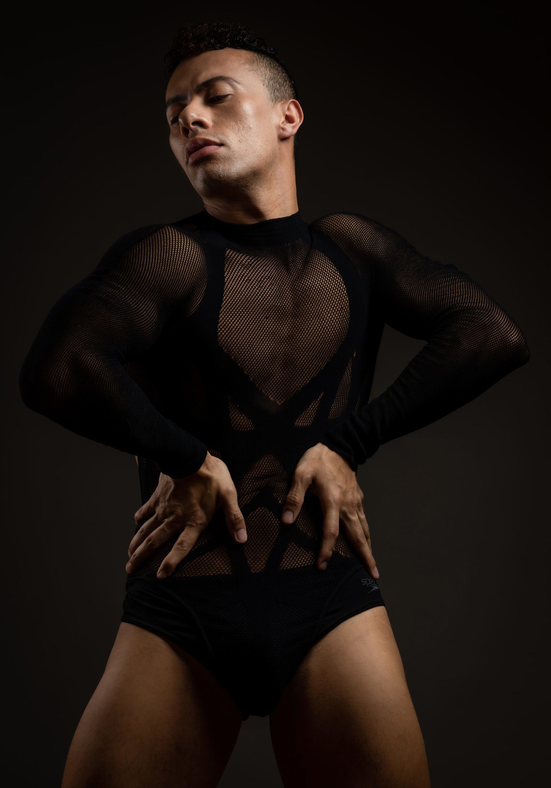 male dancer with hands on hips wearing fishnet leotard looking down