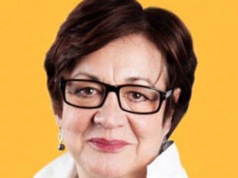 Headshot of Susannah Simons. White female with short dark hair. Wearing rectangular glasses and a white shirt on a yeallow background