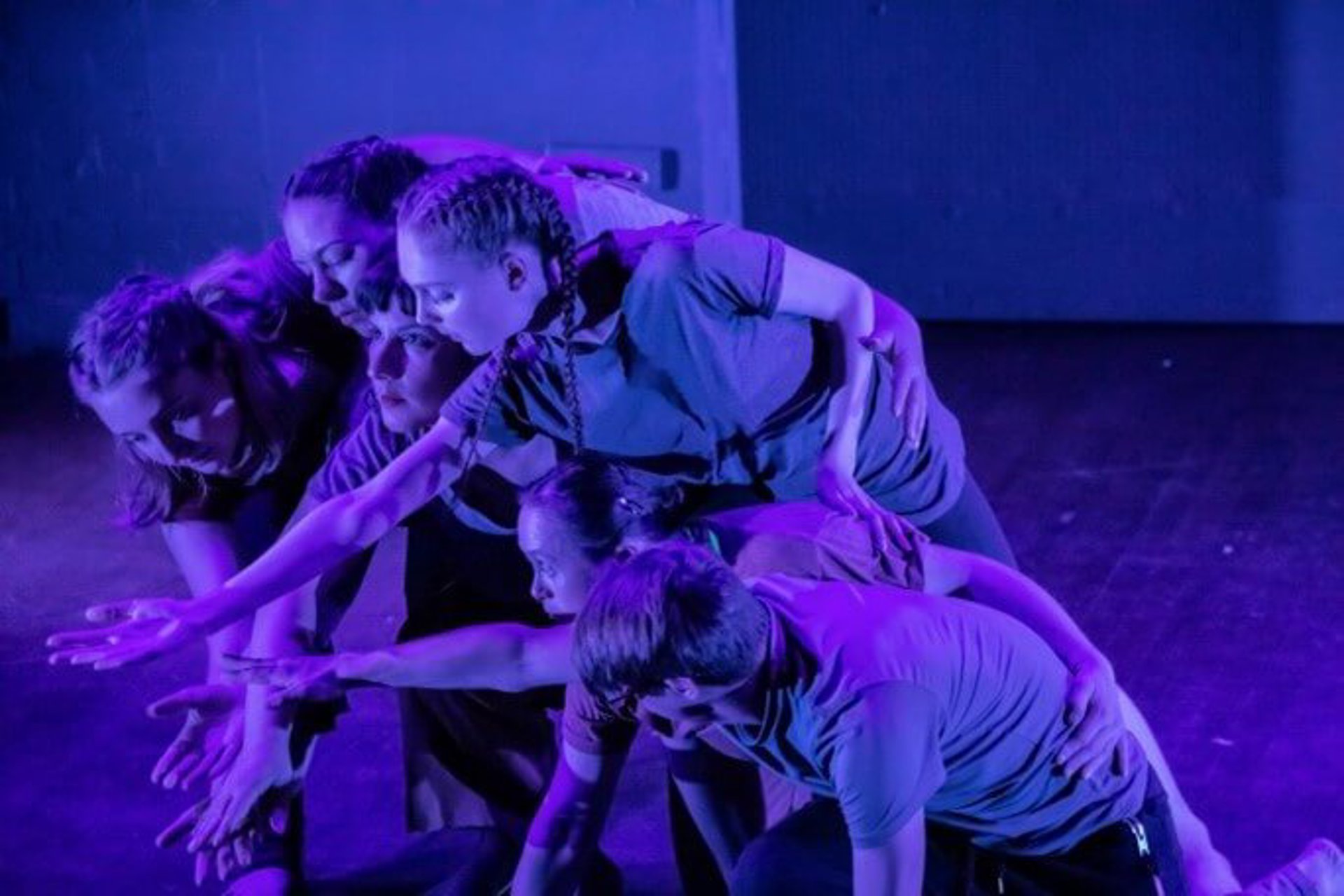 six young dancers all huddled together reaching for the same spot in the bottom left on the screen with blue lighting
