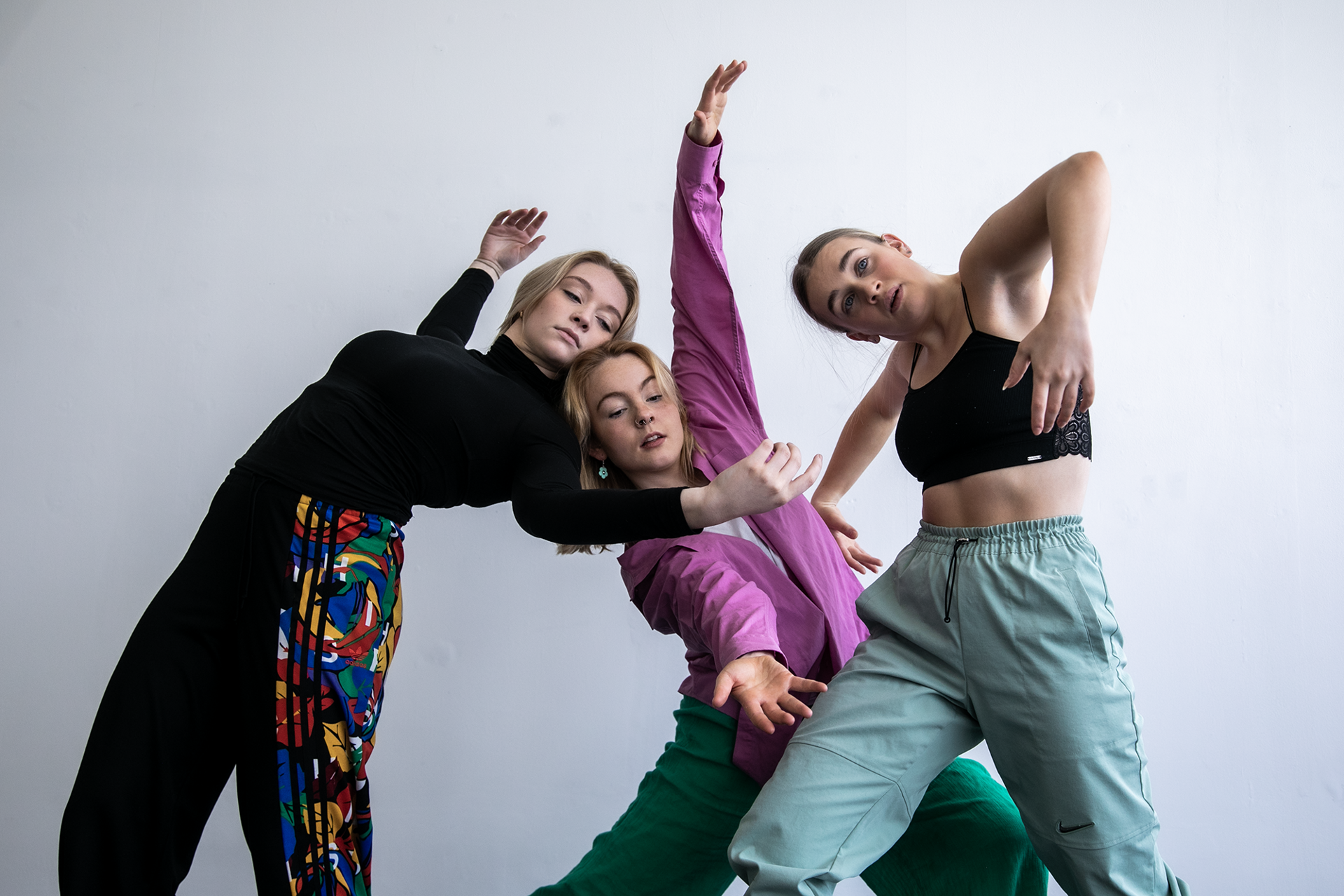 three white female dancers moving together with arms up in the air. Wearing colourful outfits against a white wall