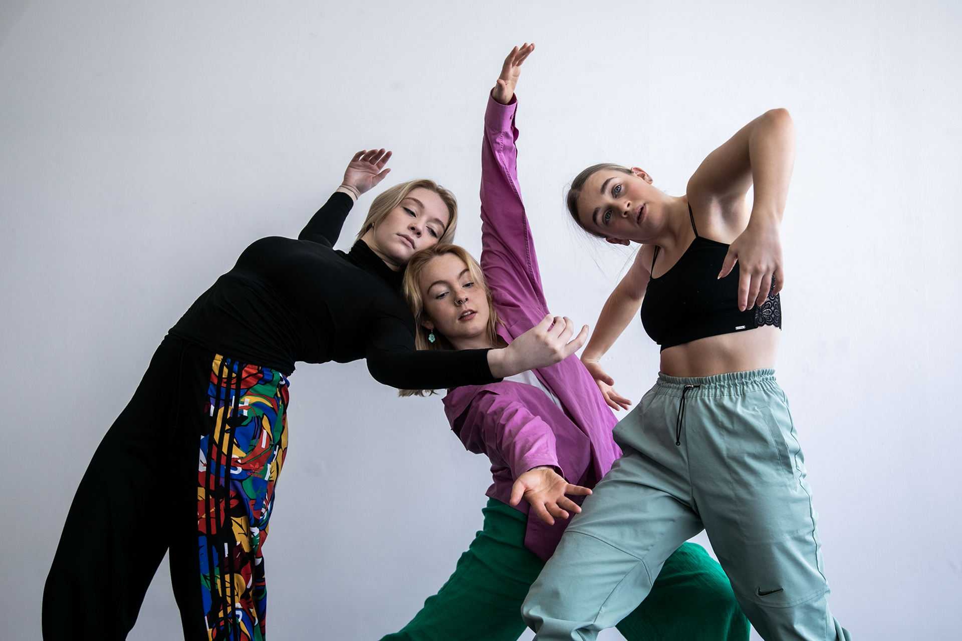 three white female dancers moving together with arms up in the air. Wearing colourful outfits against a white wall three white female dancers moving together with arms up in the air. Wearing colourful outfits against a white wall