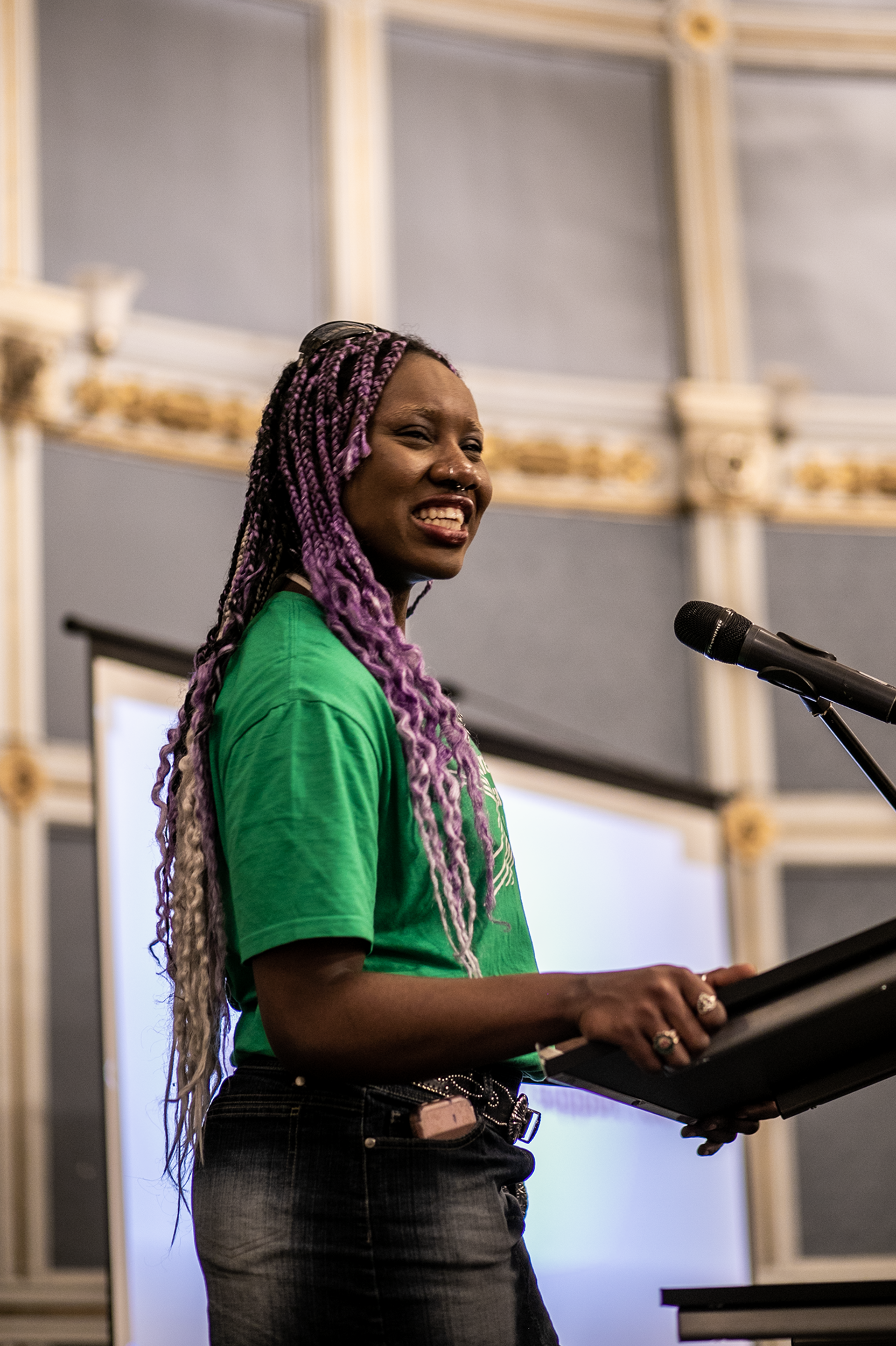 Black non binary person with purple braided hair Dance ambassador talking on stage at One Dance UK AGM wearing green tshirt Black non binary person with purple braided hair Dance ambassador talking on stage at One Dance UK AGM wearing green tshirt