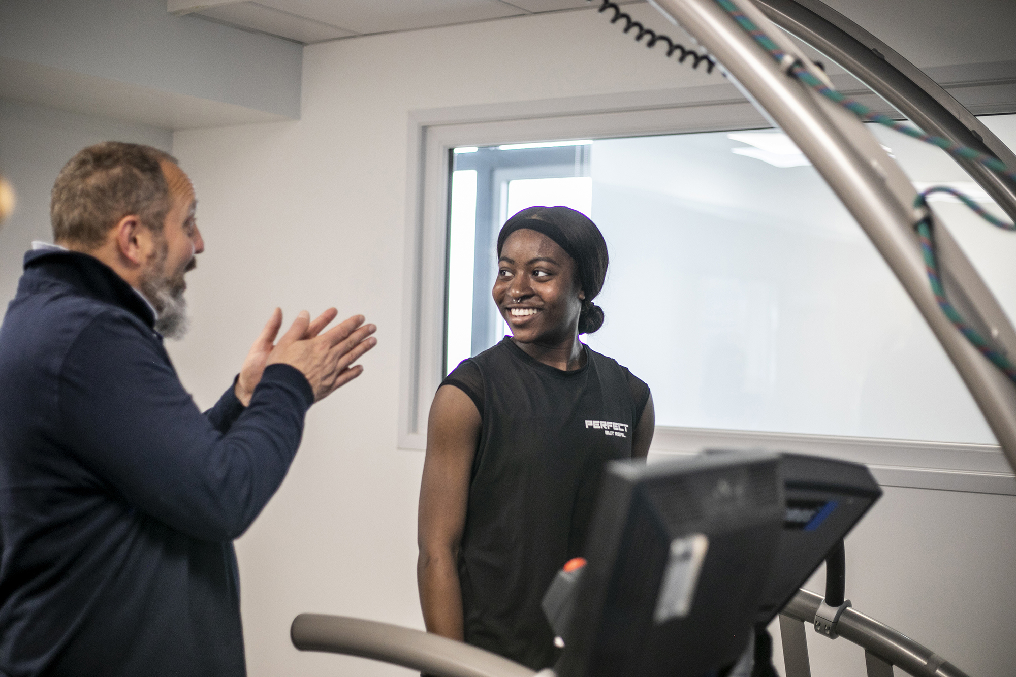 Global majority female dancer standing on a treadmill smiling whilst white male physiologist is explaining something to her. In a white laboratory setting with a window in the background