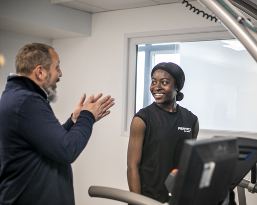 Global majority female dancer standing on a treadmill smiling whilst white male physiologist is explaining something to her. In a white laboratory setting with a window in the background