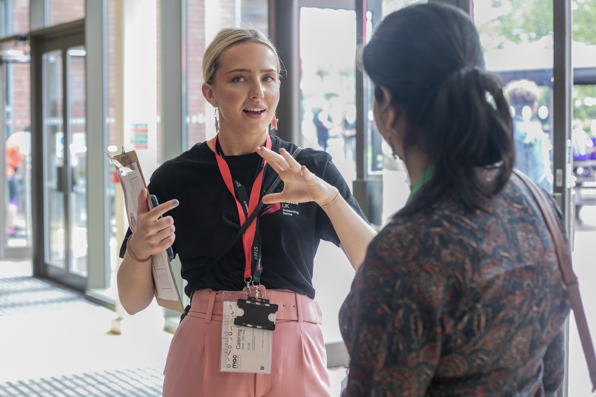 White female with tied back blonde hair One Dance UK Staff member gesturing to someone in discussion in front of glass doors