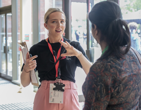 White female with tied back blonde hair One Dance UK Staff member gesturing to someone in discussion in front of glass doors White female with tied back blonde hair One Dance UK Staff member gesturing to someone in discussion in front of glass doors
