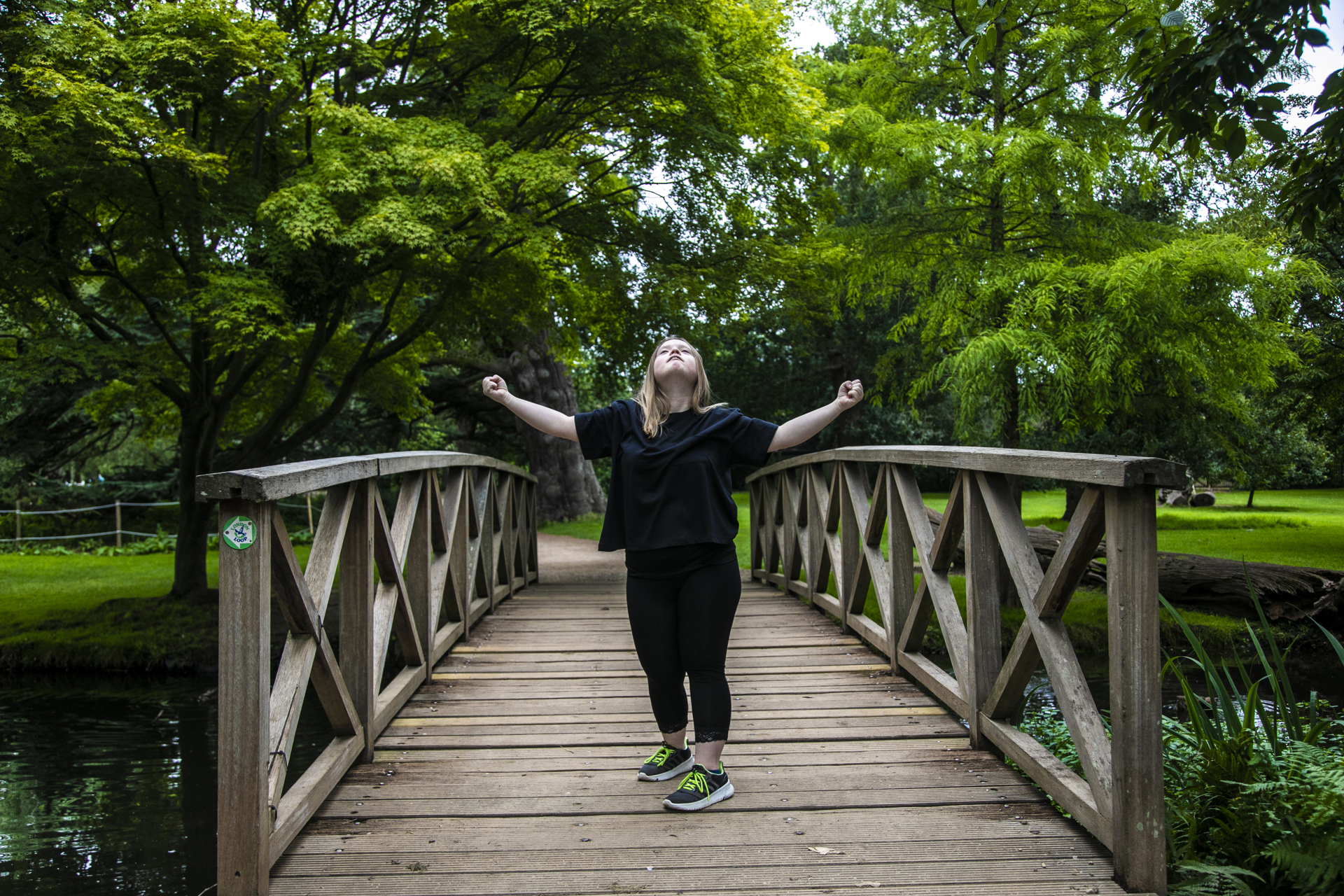 Blonde white female down's syndrome dancer wearing all black. Arms out wide in a strong stance standing in the middle of a wooden bridge with trees around. Blonde white female down's syndrome dancer wearing all black. Arms out wide in a strong stance standing in the middle of a wooden bridge with trees around.