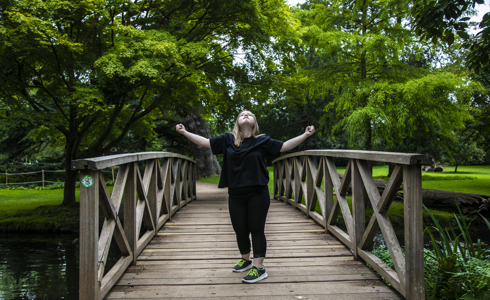 Blonde white female down's syndrome dancer wearing all black. Arms out wide in a strong stance standing in the middle of a wooden bridge with trees around.