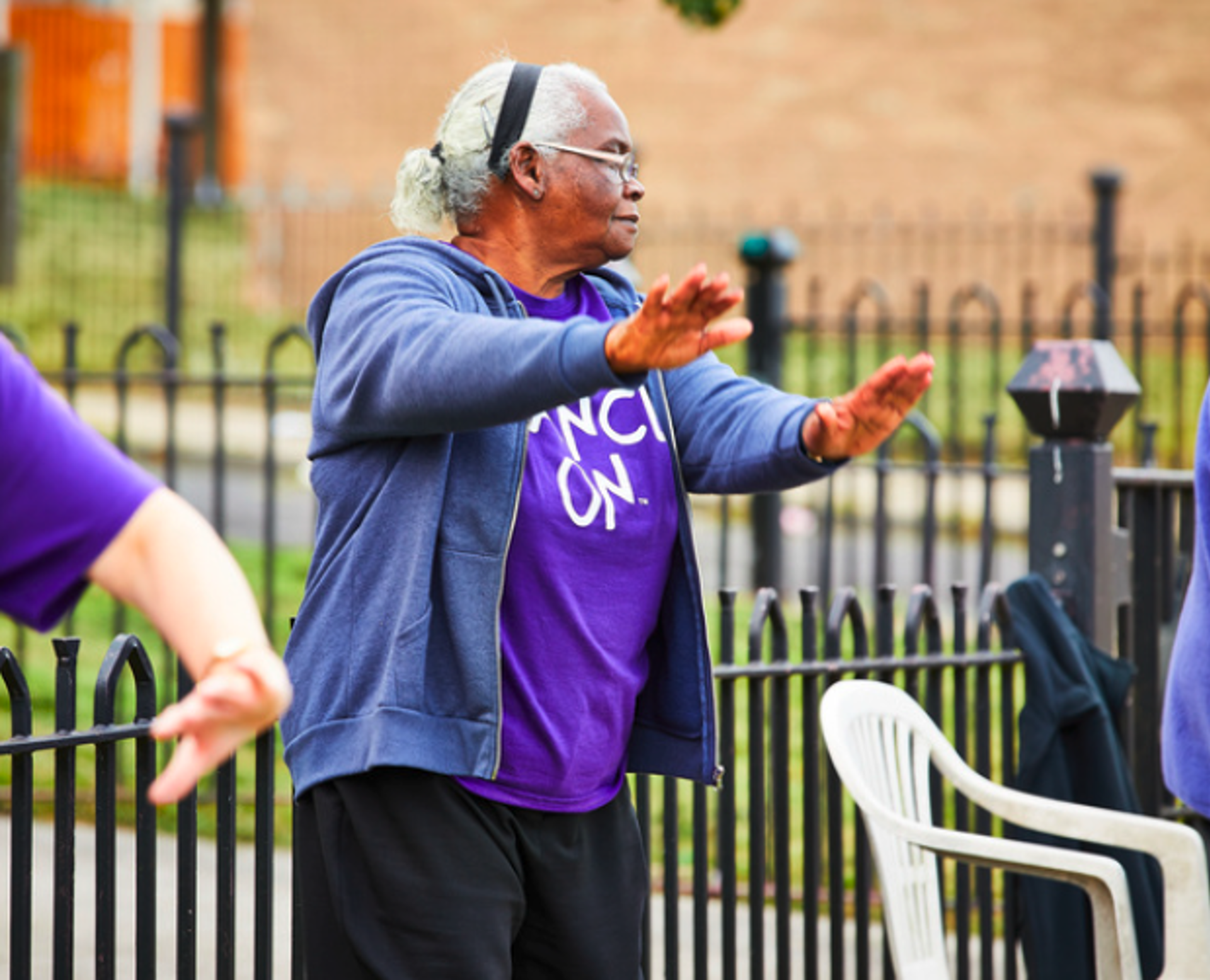 One older global majority female dancer with arms out in front of her, wearing purple top outdoors in front of black gate.