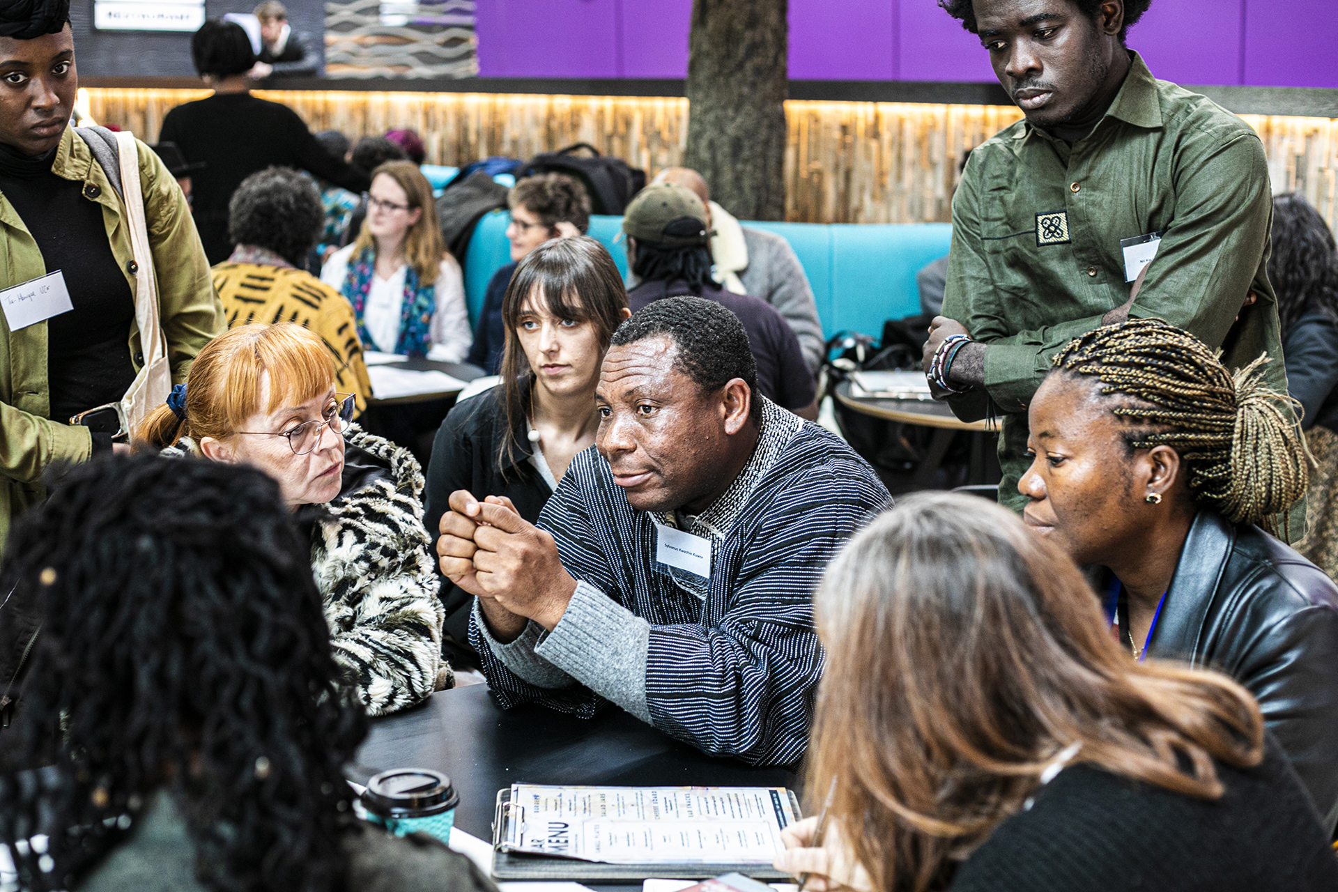 Global majority male in the centre of the image sitting at a table holding his hands together with a group of people listening to him of mixed ages, genders and ethnicities.  